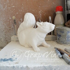 May include: White ceramic squirrel figurine with a bushy tail. The figurine is sitting on a white surface with tools and paint supplies in the background.
