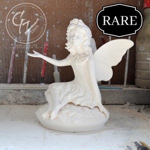 Może przedstawiać: Biała ceramiczna figurka wróżki z skrzydłami, siedząca na podstawie w kształcie liścia. Wróżka ma koronę z kwiatów i naszyjnik z kwiatów. Figurka jest oznaczona "G7 1998" i "Made in the USA".