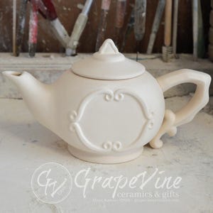 Puede incluir: Tetera de cerámica blanca con un diseño ornamentado y una tapa. La tetera tiene una asa y un pico.