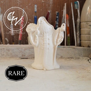 May include: White ceramic figurine of a bearded man holding a lantern and a bag of gifts. The figurine is labelled "Rare" and "Made in the USA Grapevine Ceramics & Gifts Hays KS".