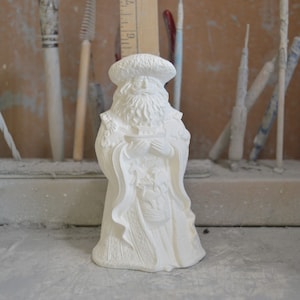 Könnte beinhalten: Weiße Keramikfigur eines Weihnachtsmanns, der ein kleines Boot in seinen Händen hält. Er trägt eine lange Robe mit einem pelzbesetzten Hut und Bart.