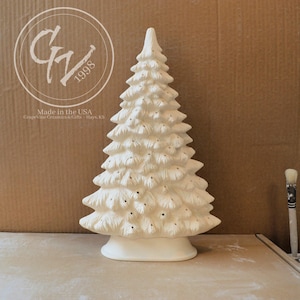 Könnte beinhalten: Eine weiße Weihnachtsbaumfigur aus Keramik mit strukturiertem, geschichtetem Zweigdesign und zahlreichen kleinen, gleichmäßig verteilten kreisförmigen Löchern auf einer glatten, ovalen Basis, wobei rechts ein Pinsel zu sehen ist.