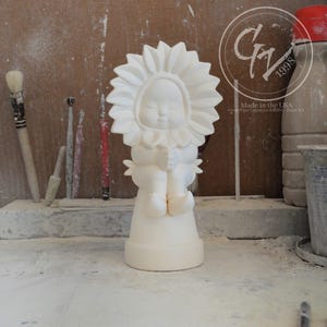 Puede incluir: Figurilla de cerámica blanca de un niño con cabeza de girasol, sentado en una maceta. La figurita está sin pintar y lista para ser decorada.