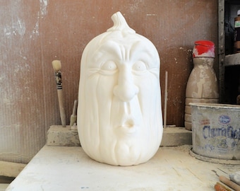 May include: White ceramic pumpkin with a carved face. The pumpkin has a wide mouth, large eyes, and a prominent nose. The pumpkin is sitting on a white surface.