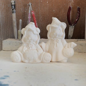 May include: Two white ceramic Halloween-themed bear figurines. One bear is wearing a witch hat and holding a pumpkin. The other bear is wearing a ghost costume and holding a trick-or-treat bag.