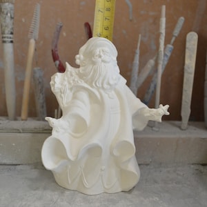 Puede incluir: Figura de cerámica blanca de Papá Noel con una larga barba blanca y un gorro rojo. Lleva una bolsa de regalos en una mano y un bastón de caramelo en la otra.