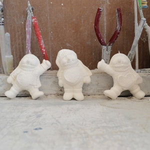 May include: Three white ceramic snowman ornaments with a heart design on their chests. The ornaments are sitting on a white surface.