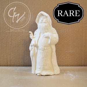 Puede incluir: Una figura de cerámica color bizcocho de Papá Noel, sosteniendo un ciervo, con una larga barba blanca y una capa con capucha. La figura tiene un cinturón y está marcada "RARE" en una etiqueta en blanco y negro. También incluye el texto "Made in the USA".