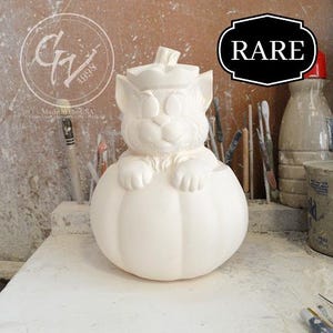 May include: A white ceramic cat figurine perched atop a pumpkin. The cat has large ears and a smiling face. The pumpkin has vertical ridges. The word "RARE" is in a black and white hexagon. The background includes art supplies.