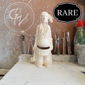 May include: White ceramic figurine of a Native American man wearing a headdress and traditional clothing. The figurine is standing on a table with a wooden surface. The figurine is labeled "RARE".