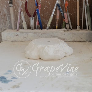 Może przedstawiać: Biała ceramiczna podstawa z detalicznym wzorem kwiatowym. Podstawa ma kształt okrągły z podniesionym środkiem i lekko nierównym brzegiem. Podstawa stoi na białej powierzchni.