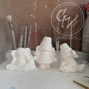May include: Three white ceramic bunny figurines with hats. The bunnies are sitting on a wooden surface. The bunnies are blank and ready to be painted.