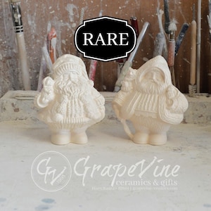 May include: Two white ceramic Santa Claus figurines, one holding a sack and the other holding a walking stick. The figurines are standing on a wooden surface. The word "RARE" is written in black letters on a black background.