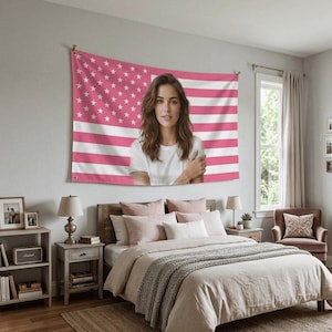 Puede incluir: Un tapiz de la bandera estadounidense rosa y blanca con la imagen de una mujer en el centro cuelga sobre una cama. El dormitorio tiene una paleta de colores neutros con una estructura de cama de madera, mesitas de noche y un sillón acogedor. La bandera está montada sobre clavijas de madera.