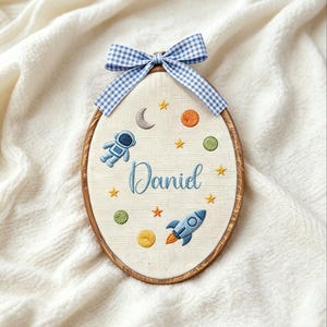 Puede incluir: Arte de aro bordado con temática espacial. El diseño incluye un astronauta, un cohete, planetas, estrellas y el nombre "Daniel" en hilo azul. Un lazo de cuadros azul y blanco adorna el aro de madera.
