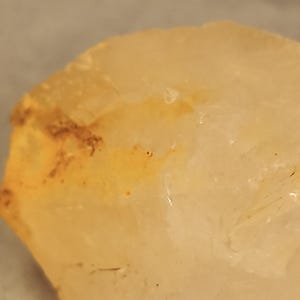 May include: Close-up of a translucent, light-yellow crystal with areas of orange and brown coloration. The crystal has a rough, uneven surface and appears to be a natural mineral specimen. The overall tone is warm and earthy.