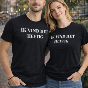 Op de afbeelding: Twee mensen dragen zwarte t-shirts met de witte tekst "IK VIND HET HEFTIG". Ze staan voor een vage achtergrond van bomen en gebouwen. Beiden dragen een blauwe spijkerbroek. De t-shirts zijn korte mouwen.