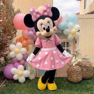 Puede incluir: Una persona con un disfraz de Minnie Mouse, vestida con un vestido rosa con lunares, guantes blancos y zapatos amarillos. Un gran lazo rosa con lunares blancos está en la cabeza. El fondo incluye globos y flores decorativas.