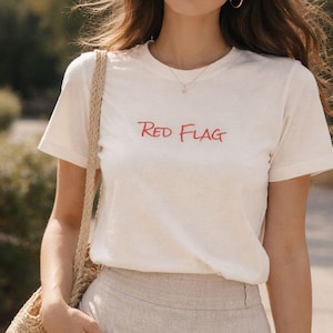 以下が含まれることがあります： 赤色の「Red Flag」の文字が入ったクリーム色のTシャツ。重ねフリルデザインのベージュのリネンスカートと合わせています。カジュアルウェアに適した、織りのショルダーバッグがコーディネートを完成させています。