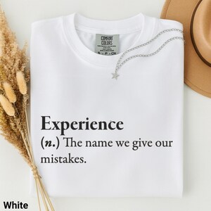 May include: White t-shirt with the text "Experience (n.) The name we give our mistakes." A silver necklace with a star pendant and a tan hat are on the shirt. The shirt has a Comfort Colors tag.