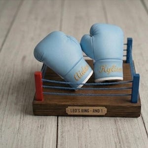 Puede incluir: Un par de guantes de boxeo azul claro con letras doradas, descansando sobre un ring de boxeo de madera en miniatura. El ring tiene postes rojos y azules y la base dice "Leo's Ring - Rnd 1". Los guantes están personalizados.