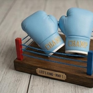 May include: A miniature boxing ring with light blue boxing gloves. The gloves have "CHAMP" embroidered in gold. The ring is made of wood with blue and red accents. The base has a plaque that reads "LEO'S RING - RND 1".