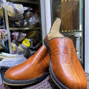 Könnte beinhalten: Ein Paar braune Leder-Slipper mit dunkler Sohle. Die Schuhe haben eine strukturierte Oberfläche mit einem genähten Design. Die Innenseite ist hellbraun. Die Schuhe werden auf einer strukturierten Oberfläche präsentiert.