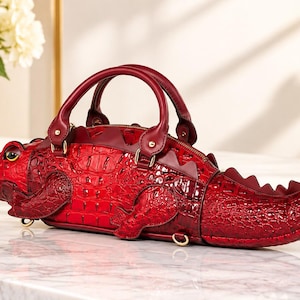 May include: A deep red handbag shaped like a crocodile with a textured, embossed leather-like finish. The bag features a prominent snout with a green eye, two top handles, a zippered main compartment, and a curved tail extending to the right.