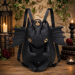 May include: A black backpack shaped like a creature with large, pointed ears, two prominent horns, and wide, amber-coloured eyes. The bag features two extended, bat-like wings on the sides, two small arms resting on the front, and two shoulder straps.