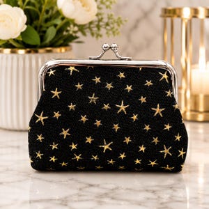 May include: A black coin purse featuring a gold star pattern across the fabric. The purse has a silver-toned metal frame with a kiss-lock clasp closure at the top. The exterior fabric has a textured weave, and the metal hardware is polished.