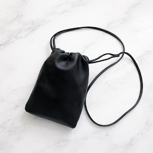 Può includere: Una borsa nera in pelle con coulisse e una tracolla lunga e sottile. La borsa è rettangolare con angoli arrotondati e una parte superiore arricciata. La tracolla è attaccata alla borsa con un nodo su ogni lato.