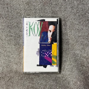 Könnte beinhalten: Eine Vintage-Kassette mit dem Album "Lucky Man" von Dave Koz. Das Cover zeigt ein Porträt eines Mannes, den Namen des Künstlers und ein farbenfrohes Design mit Würfeln. Das Band befindet sich in einer transparenten Kunststoffhülle.