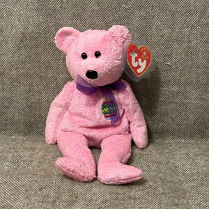 May include: A pink plush bear with a purple ribbon and an Easter egg design. The bear has black eyes and a black nose. A Ty tag is attached to the ear. The bear is sitting on a textured surface.