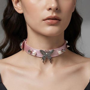 May include: A light pink choker necklace featuring a central silver-toned butterfly charm, alternating white faux pearls, and metallic cone-shaped spikes along the band. The accessory is worn around the neck, showcasing a smooth satin-like finish.