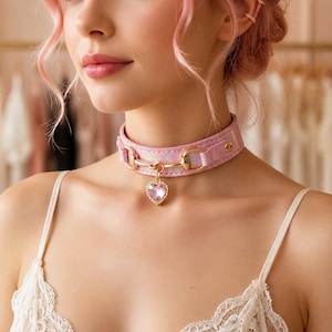 Puede incluir: Una gargantilla de color rosa iridiscente con un herraje metálico dorado en forma de bocado en el centro. Un colgante rosa en forma de corazón con facetas y borde dorado cuelga del centro del herraje metálico de la gargantilla.