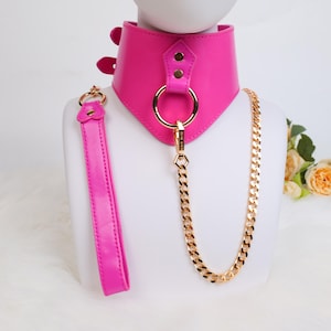 Puede incluir: Un conjunto de collar y correa rosa brillante. El collar es ancho con herrajes dorados, incluyendo un gran anillo y una cadena. La correa es rosa a juego con un asa de bucle. El conjunto se exhibe en una cabeza de maniquí blanca.
