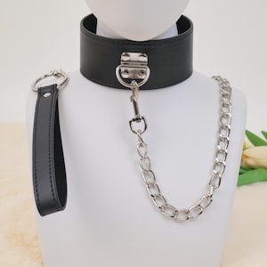 Puede incluir: Un collar de cuero negro con una banda ancha y una correa de cadena plateada. El collar tiene un cierre de metal y un asa a juego. Los artículos se exhiben en una cabeza de maniquí blanca.