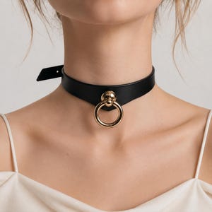 Puede incluir: Un collar gargantilla negro con un colgante de anillo dorado. La gargantilla está hecha de un material liso y oscuro y se abrocha con una hebilla. El anillo dorado es una característica destacada, que añade un toque de elegancia al diseño.