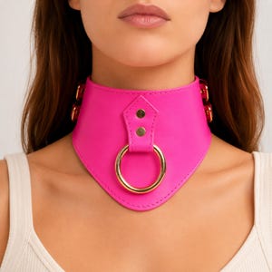 Puede incluir: Un collar gargantilla rosa brillante con un anillo y herrajes dorados. El collar está hecho de un material liso y presenta un diseño frontal puntiagudo. El anillo dorado está centrado en la parte delantera del collar.