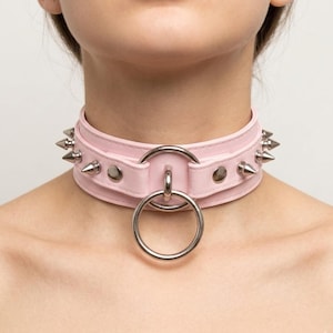 Puede incluir: Un collar choker rosa claro con púas plateadas y un gran anillo de metal. El collar está hecho de un material liso y tiene un cierre de hebilla. Las púas están espaciadas uniformemente.