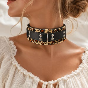 Puede incluir: Un collar de cuero negro con una banda con estampado de leopardo. El collar presenta remaches plateados y un anillo de metal plateado. La banda con estampado de leopardo tiene un fondo beige con manchas marrones y negras.