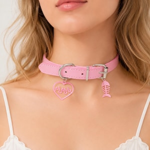 Puede incluir: Un collar choker rosa con herrajes plateados. El collar presenta dos colgantes: un corazón con diseño de llama y una espina de pescado. El collar está hecho de un material liso.