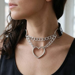 Puede incluir: Una gargantilla de cuero negro con un diseño de cadena de metal plateado. El accesorio incluye dos cadenas plateadas superpuestas conectadas a un anillo central en forma de corazón. La gargantilla se asegura con una hebilla de metal plateado.