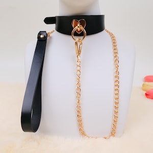 Puede incluir: Collar de cuero negro con un detalle dorado en forma de corazón y una correa de cadena a juego. El collar tiene un cierre de hebilla y un asa para la correa.