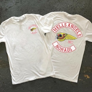 May include: Two white t-shirts with the "Hells Angels Nomads" logo printed on them. The logo features a winged skull and the words "Hells Angels" and "Nomads" in red. The shirts are on a grey surface.