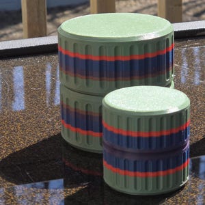 May include: Three green cylindrical containers with ribbed sides and flat tops. Each container has two horizontal stripes: a red stripe between two blue stripes. The containers are stacked on a reflective surface.