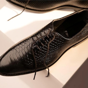 Könnte beinhalten: Schwarzer Lederschuh mit einem strukturierten, schuppenartigen Muster. Der Schuh hat eine spitze Zehenpartie und ist mit dunkelbraunen Schnürsenkeln geschnürt. Der Schuh ist vor einem weißen Hintergrund platziert.