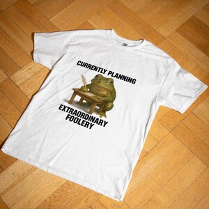 Può includere: Una t-shirt bianca a maniche corte con la grafica di una rana verde con occhiali, seduta a una scrivania di legno con una penna d'oca. Il testo sopra recita CURRENTLY PLANNING e il testo sotto recita EXTRAORDINARY FOOLERY.