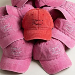 Puede incluir: Una colección de gorras de béisbol rosas y rojas. Una gorra roja presenta el texto "Wife on a Mission! THE BRIDE" en escritura negra. Otras gorras son rosas y tienen "Support Squad! THE CREW" en escritura blanca.