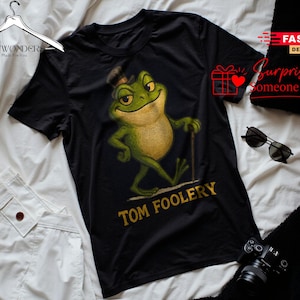 May include: A black short-sleeve t-shirt featuring a graphic of a green frog wearing a top hat and holding a cane. The frog is depicted in a walking pose with a smirk. Below the frog, the text TOM FOOLERY is printed in a bold yellow font.